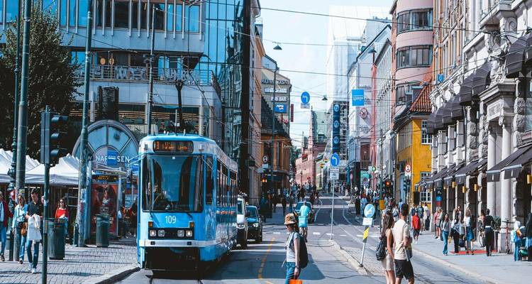 Calle concurrida de Oslo con un tranvía azul, cables aéreos y peatones bajo un cielo despejado.