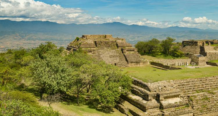 Alte Steinstrukturen von Monte Albán auf einem grünen Hügel mit entfernten Bergen.