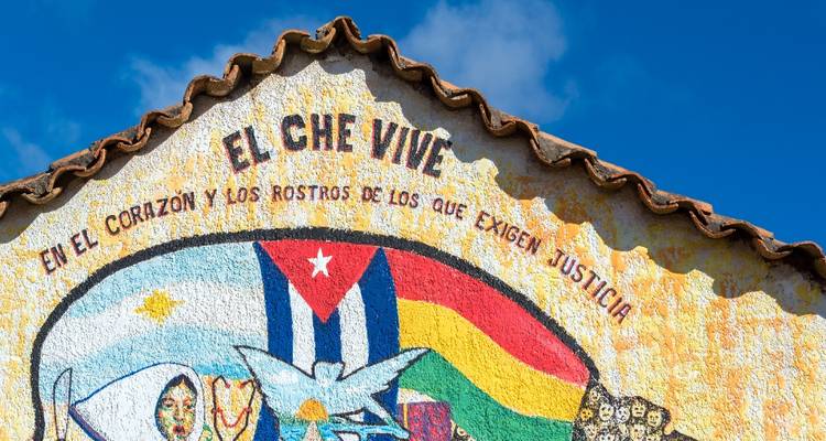 Mural colorido en una pared rústica proclamando 'EL CHE VIVE' con banderas y símbolos