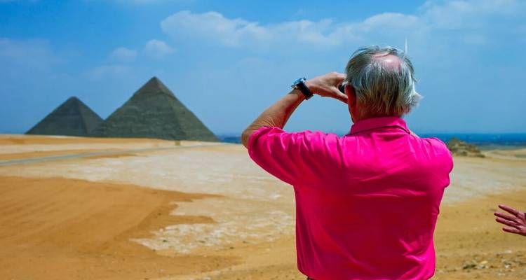 Een oudere reiziger in een felroze shirt fotografeert twee verre pyramides aan de overkant van de woestijn.