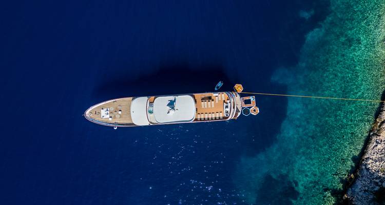 Perfectly centered vertical drone shot of the yacht anchored beside vivid turquoise shallows and deep navy water.