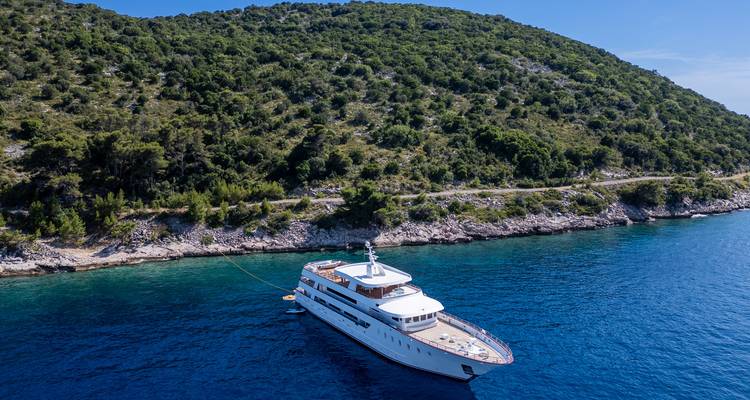 Side aerial of yacht moored along forested rocky shoreline with clear water showing different blue tones.