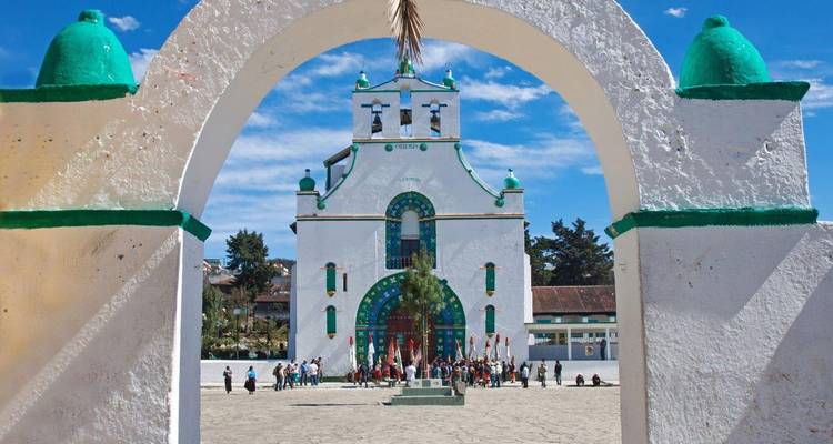 Witte kerk van San Juan Chamula met groene accenten, omlijst door een adobeboog tegen een blauwe hemel.