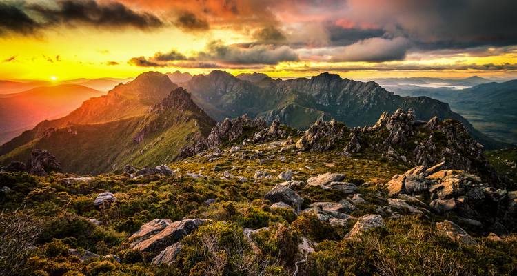 Spektakulärer Sonnenuntergang über gezackten Bergkämmen mit leuchtenden Wolken und tasmanischer Wildnis darunter.
