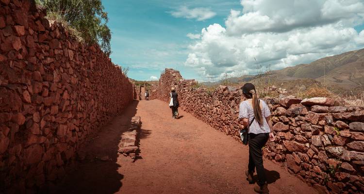 Les voyageurs se promènent le long d'un couloir de terre rouge bordé par des murs de pierre anciens sous des nuages cotonneux.