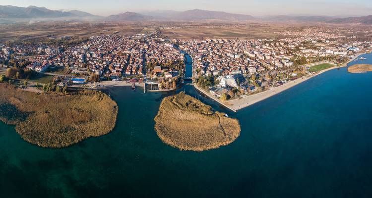 Aerial panorama of a lakeside town with a canal flowing into a wide blue lake and reed-covered islets, backed by distant mountains.