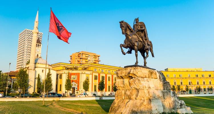 Equestrian statue of a national hero beside the Albanian flag in a sunny city square with historic and modern buildings.