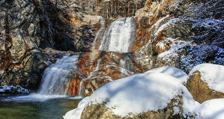 Multi-tiered waterfall cascading over reddish rocks, partially covered in fresh snow and surrounded by winter forest.