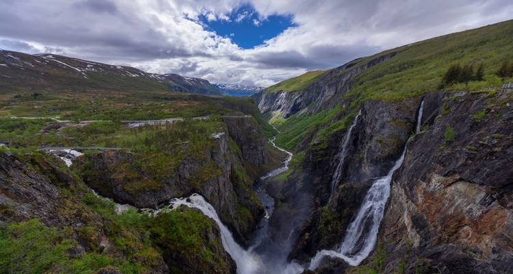 Dramatic twin-plunge Vøringfossen waterfalls cascade into a deep gorge amid steep green cliffs and distant snowy peaks.