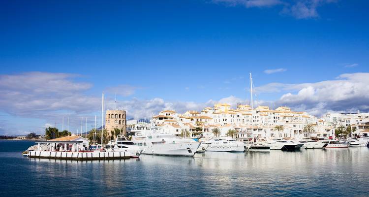 Luxe jachten afgemeerd in de Puerto Banus-marina met witte Andalusische gebouwen en blauwe lucht.