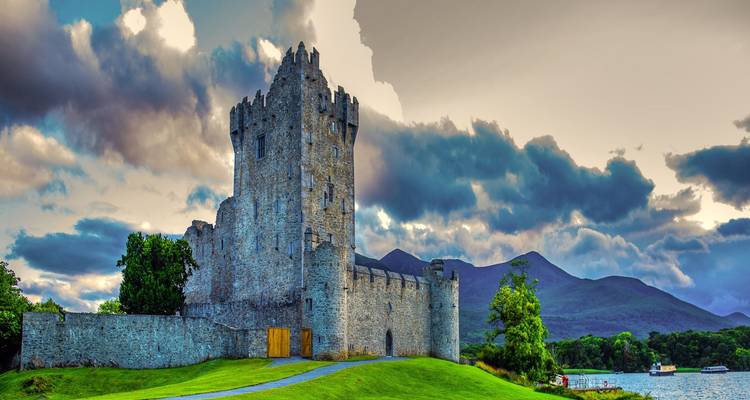 Stone Ross Castle stands on a grassy knoll beside a tranquil lake backed by misty mountains and dramatic clouds.