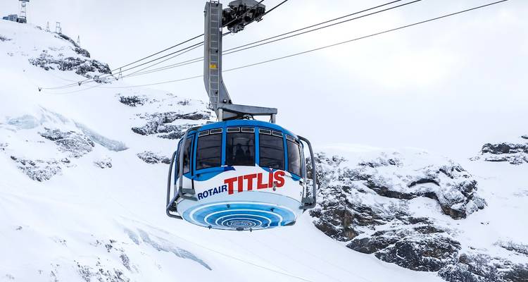 Blue rotating Titlis cable car glides above snowy alpine cliffs and glaciers on an overcast winter day.