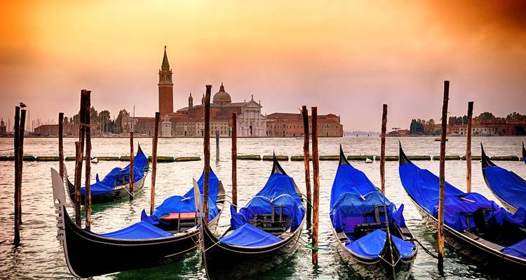 Row of blue-covered gondolas bob in front of Venice skyline at warm orange sunset light.