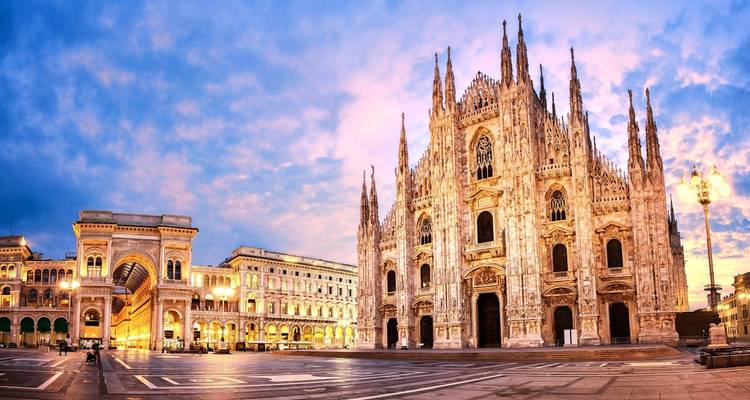 Gothic spires of the Milan Duomo glow in soft evening light beside the grand Galleria arcade on a wide plaza.