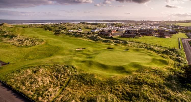 Photographie aérienne d'un parcours de golf vallonné en bord de mer à côté d'une ville côtière sous une lumière douce du soir.