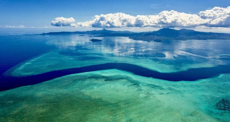 Stunning aerial of vivid blue reefs, islands and distant mountains beneath a bright sky.