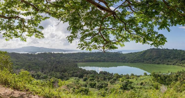 Ground-level panorama of a crater lake framed by tree branches with sea and hills beyond.