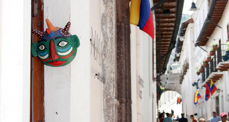 Bunte traditionelle Maske, die neben einer engen Kolonialstraße hängt, die mit ecuadorianischen Flaggen gesäumt ist