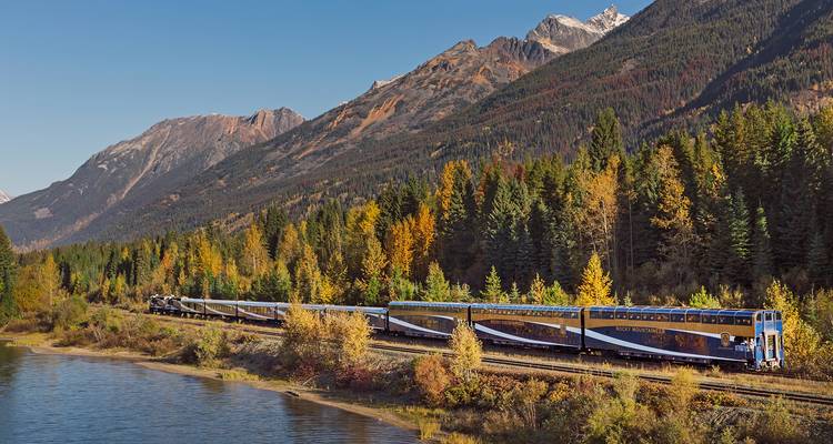 Luxe koepelwagontrein die langs een rivier slingert onder gouden herfstbossen en ruige bergtoppen.