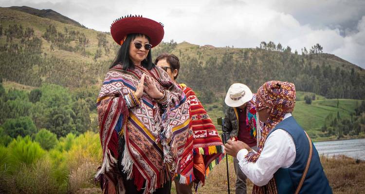 Una mujer con sombrero rojo y poncho sonríe mientras el chamán presenta una ofrenda con la cordillera de fondo.