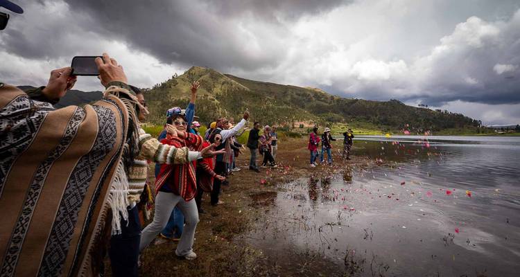 Los participantes lanzan pétalos coloridos en un lago reflectante bajo un cielo nublado y dramático.