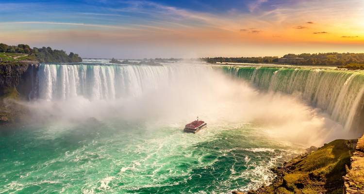 Panoramisch zonsondergangzicht op de Niagarawatervallen met rondvaartboot in turquoise water omgeven door opstijgende nevel.