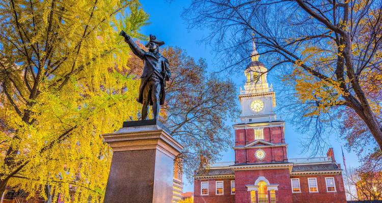 Bronzen standbeeld op een lommerrijke plein wijst naar de iconische klokkentoren van Independence Hall op een frisse herfstdag.