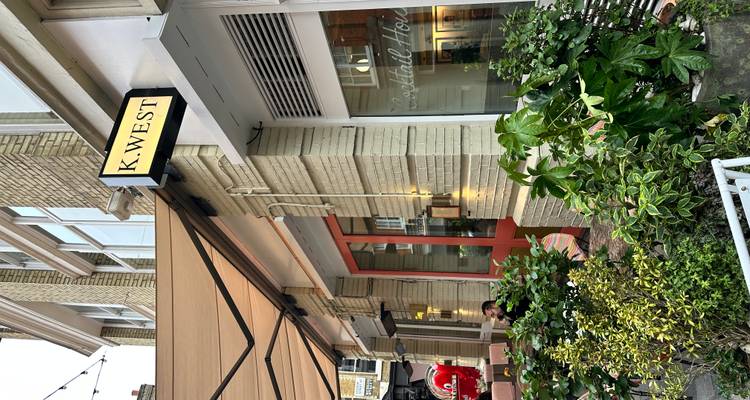 Straatgezicht van caféterras met geel 'K.WEST' bord en potplanten.