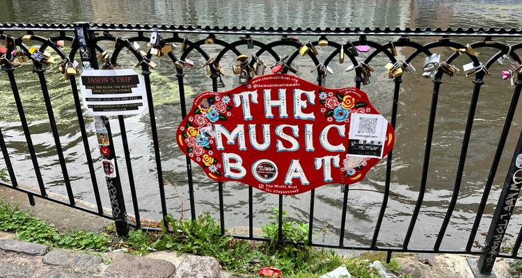 Decoratief ijzeren hek langs het kanaal met een felrood bord met 'The Music Boat' erop en veel liefdesslotjes.