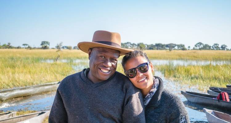 Un couple souriant pose près de pirogues mokoro sur une rive herbeuse du delta.