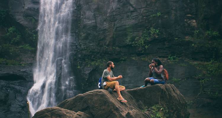 Dos viajeros sentados en una saliente rocosa charlando junto a una cascada alta en el bosque.