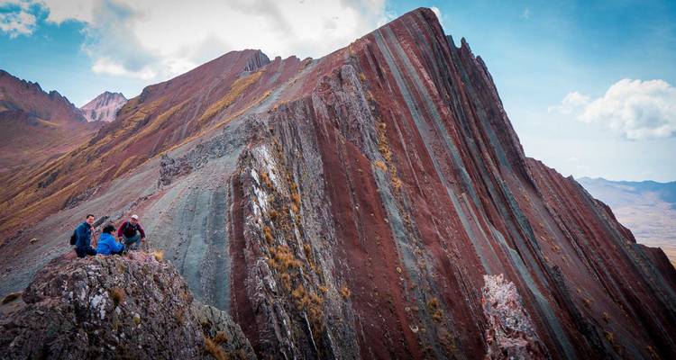 Vista amplia de una cresta multicolor y empinada con un pequeño grupo de excursionistas descansando en las rocas del primer plano bajo un cielo azul.
