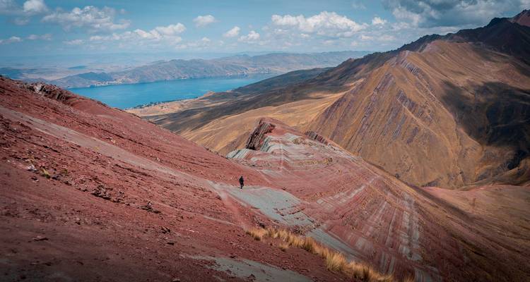 Vista panorámica de una ladera montañosa con rayas rojo y turquesa que desciende hacia un lago azul distante y una meseta.