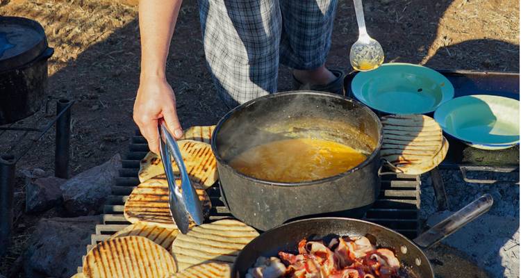 Gros plan du cuisinier du camp retournant du pain plat grillé et du bacon crépitant à côté d'une marmite de ragoût.
