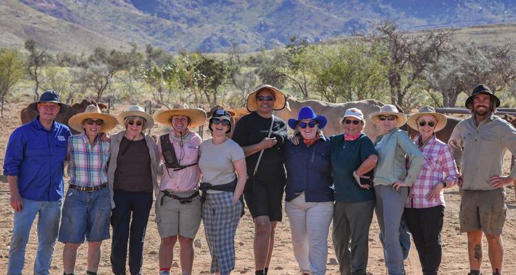 Un groupe de touristes souriants pose ensemble dans une station d'élevage poussiéreuse avec des montagnes en arrière-plan.
