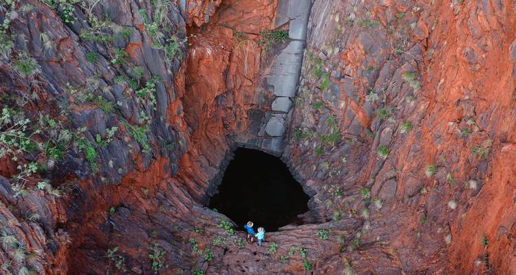 Deux explorateurs se tiennent à l'entrée d'un profond et étroit gouffre de roche rouge menant à un bassin obscur.