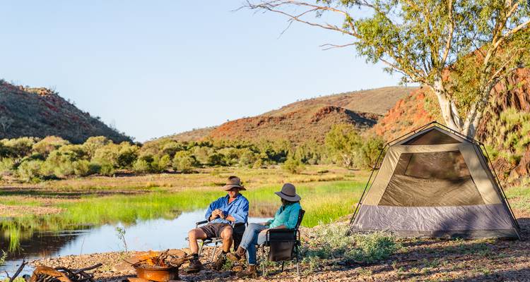 Un couple se détend près d'un feu de camp et d'une tente près d'un point d'eau réfléchissant au milieu de collines rouges.