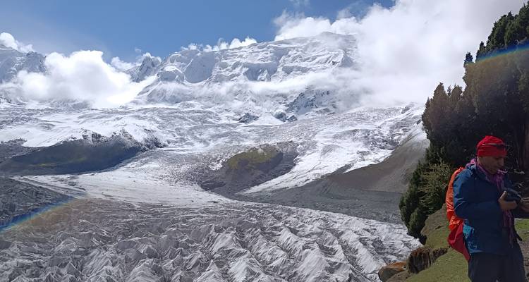 Un randonneur vêtu d'équipement voyant se tient à côté d'un glacier massif avec des sommets enneigés imposants s'élevant dans des nuages épars.