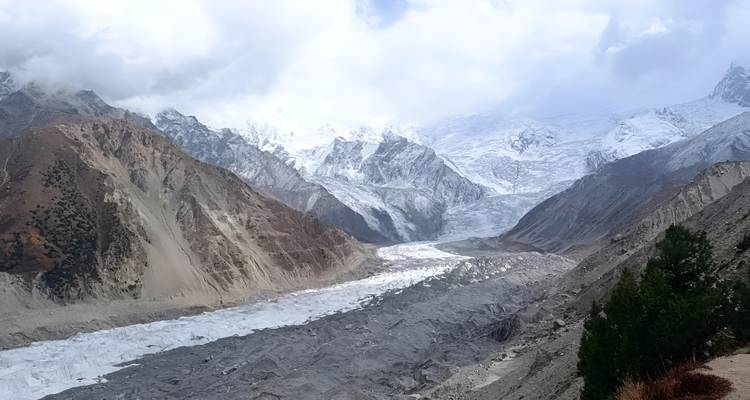 Un glacier sinueux recouvert de débris s'écoule à travers des pentes abruptes et stériles de couleur brune vers des sommets glacés lointains.