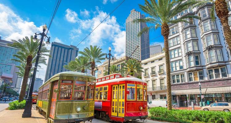 Historic streetcars in red and green glide along palm-lined tracks in downtown New Orleans on a sunny day.