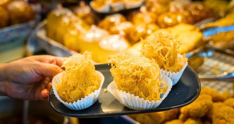 Close-up of a hand holding Thai crispy golden desserts on a market tray.