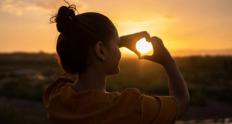 Silhouette d'une femme formant un coeur avec ses mains autour du soleil couchant sur fond de désert.