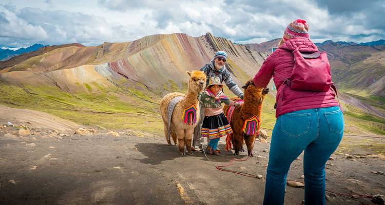 Toeristen die een kind in traditionele kleding fotograferen met alpaca's voor de gestreepte hellingen van Rainbow Mountain.