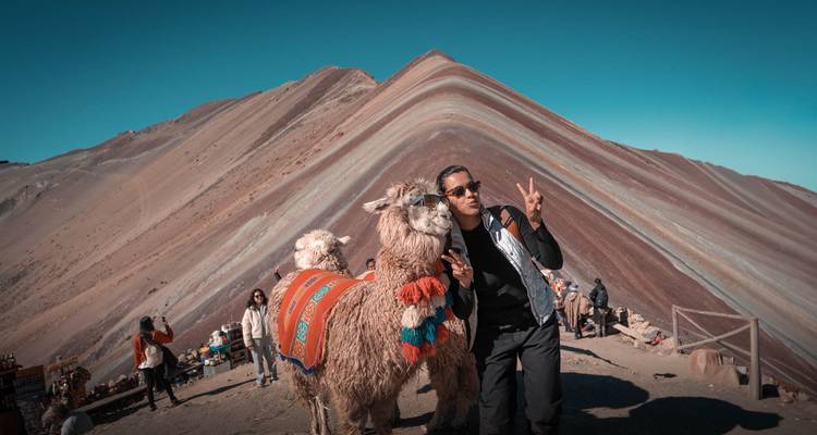Een reiziger poseert met een versierde lama terwijl hij een vredesteken maakt bij Rainbow Mountain.