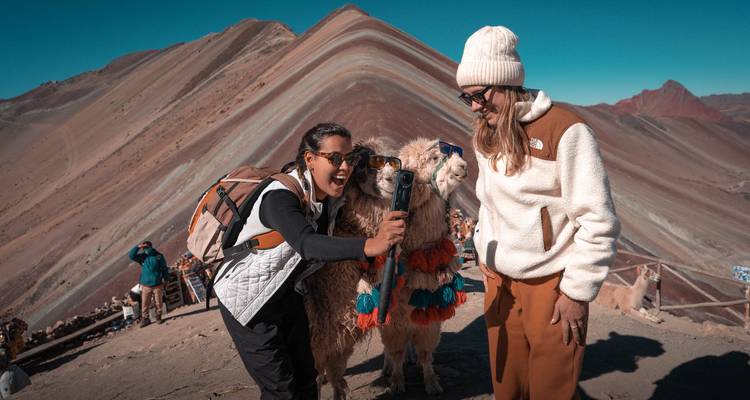 Twee vrouwen lachen terwijl ze een vriendelijke lama filmen voor de gestreepte helling van Rainbow Mountain.