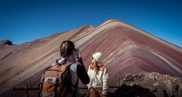 Een wandelaar fotografeert een vriend die poseert met de veelkleurige kam van Rainbow Mountain op de achtergrond.