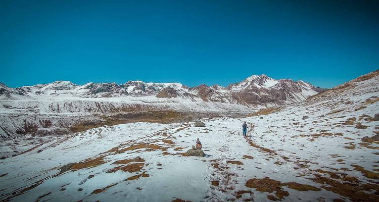 Twee wandelaars lopen over een besneeuwde bergpad onder een helderblauwe hemel en ruige toppen.