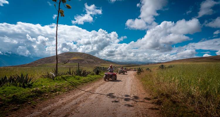 Rijders die op quad bikes over een zandweg rijden door open landbouwgebied met een lage groene berg en dramatische wolken op de achtergrond