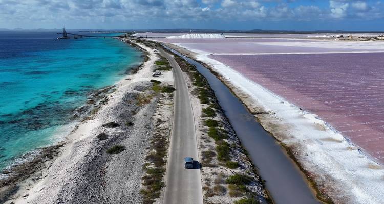 Route côtière s'étendant entre une mer turquoise et des marais salants rose-blanc sous un ciel partiellement nuageux.