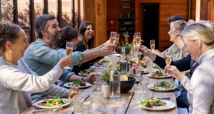 Des amis lèvent leurs verres dans une salle à manger chaleureuse en bois, partageant un repas copieux autour d'une longue table rustique remplie de plats frais.